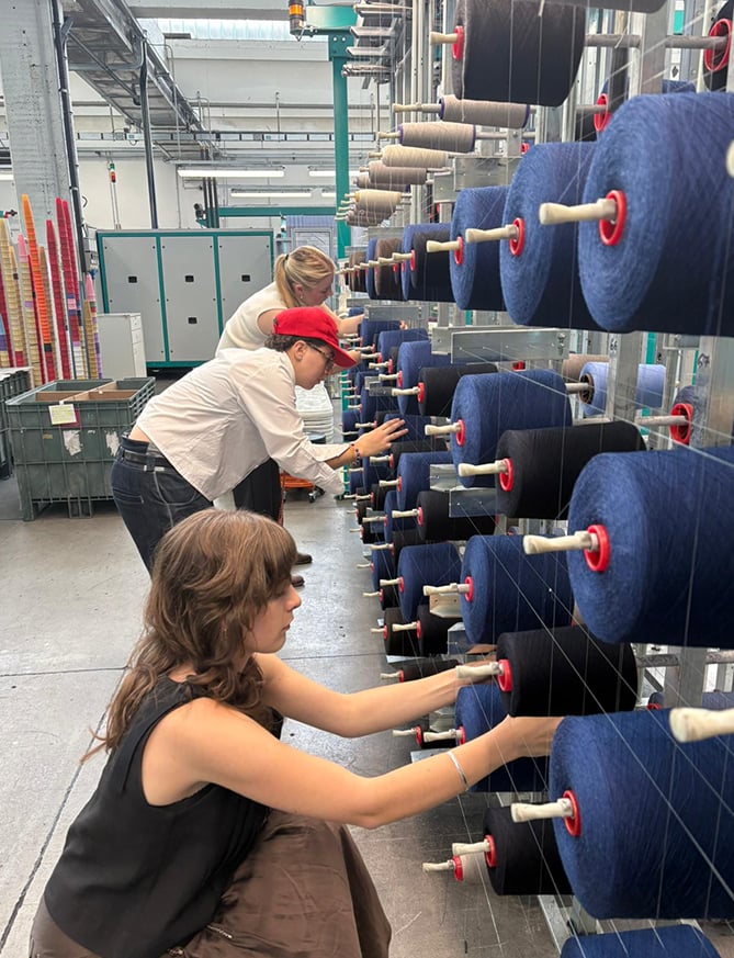 Three people load blue and black yarn cones onto a large industrial creel in a textile factory.