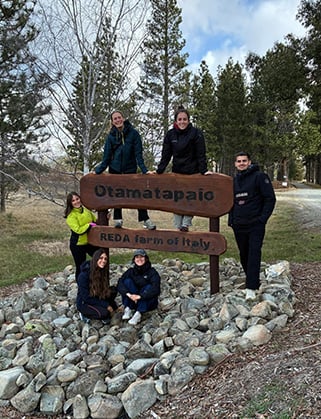Five people pose on a rock pile around a wooden sign reading "Otamatapaio – REDA farm of Italy"