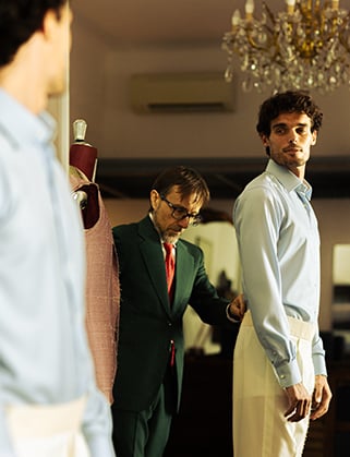 Tailor in a green suit adjusts the jacket of a man in a light blue shirt and cream trousers in a fitting room