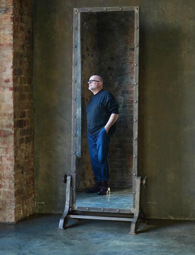 Bald bearded man with glasses, in dark sweater and blue trousers, stands in a large standing mirror in a brick room