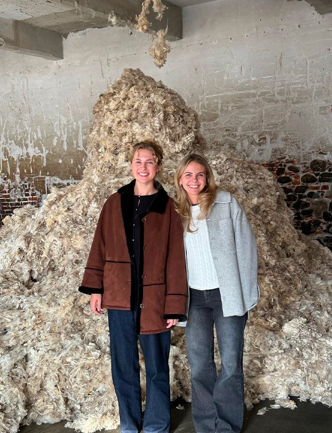 Two smiling women stand in front of a large pile of raw wool in a rustic brick-walled room