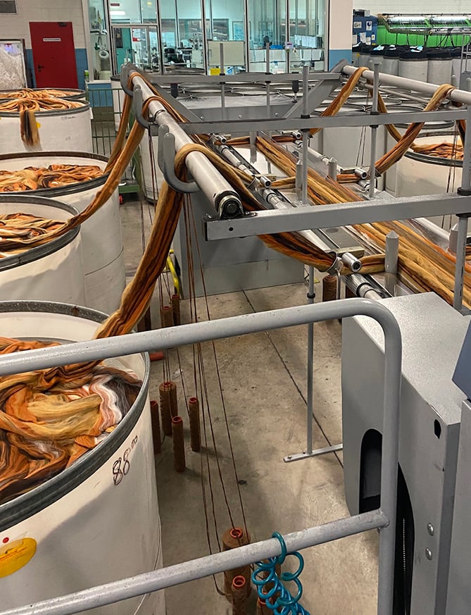 Orange and brown wool slivers run from large white cans through overhead rollers in a textile mill.