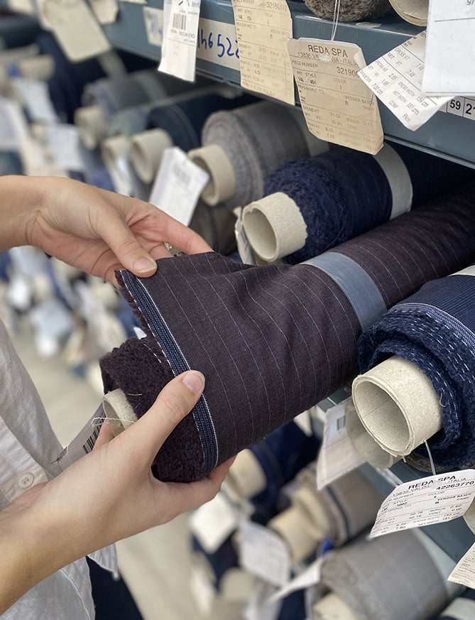 Hands examine a rolled dark purple pinstriped wool fabric on a shelf of Reda SPA fabric rolls