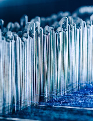 Close-up of rows of thin metal heddles with hooked tops on a loom above blue woven fabric
