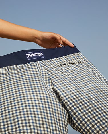 Hand holding the navy waistband of gingham shorts with a Vilebrequin label, against a clear blue sky