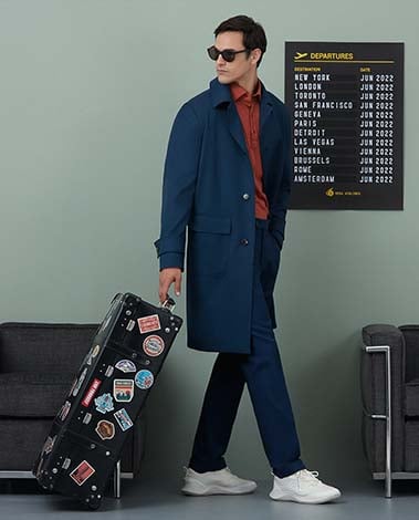 Man in navy coat and sunglasses pulls a sticker-covered suitcase past a Departures board