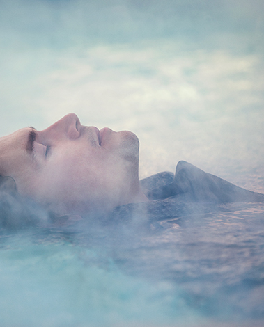 Person with closed eyes floating on their back in misty, pale blue water, face turned upward