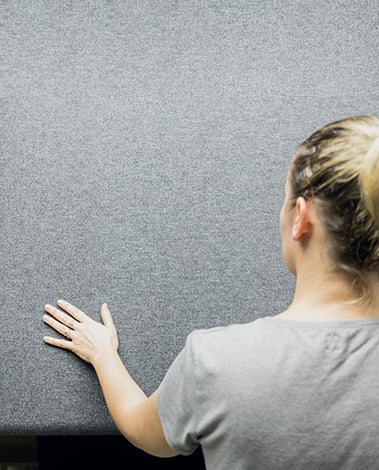 Woman seen from behind rests her hand on a gray felt-like textile wall panel