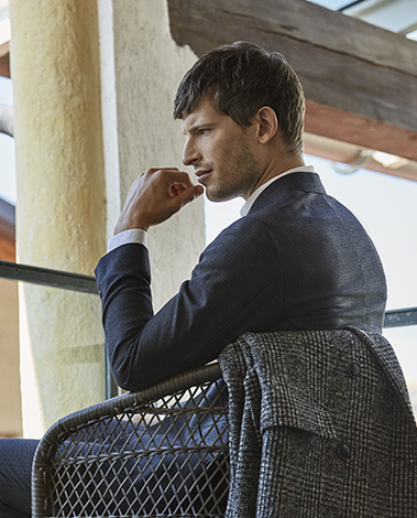 Man in a navy wool jacket sits pensively on a woven chair, a gray checked coat draped over its back
