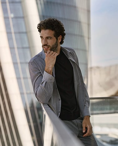 Man with curly hair leaning on a glass railing, wearing a gray wool-look shirt jacket over a black tee