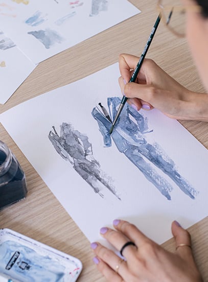 Woman painting watercolor sketches of suit-clad figures in blue and gray on paper at a wooden desk