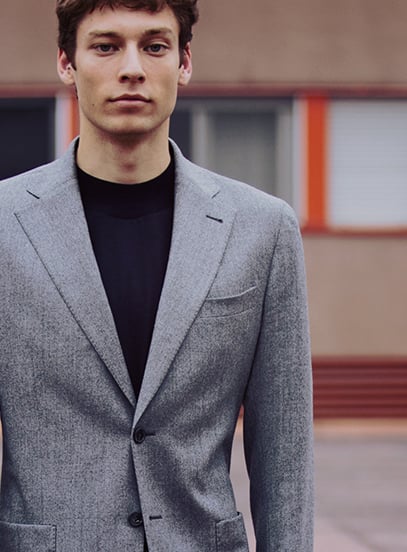 Man in a grey wool blazer over a black crew-neck sweater, standing outdoors before a building
