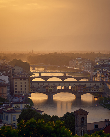 Ponte Vecchio and other bridges over the Arno at golden sunset, with rooftops and a stone tower in Florence