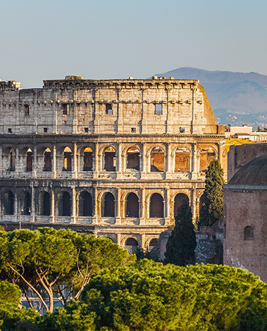 The Colosseum in Rome at golden hour, seen above green pine trees with hills in the distance