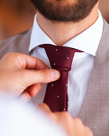 Hand adjusting a burgundy polka-dot tie on a man in a white shirt and gray vest