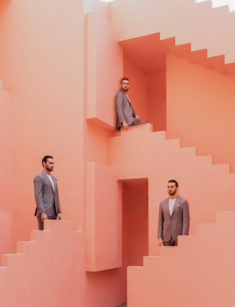 Three men in gray suits stand and sit on pink stairs and walls of a geometric salmon-colored building.