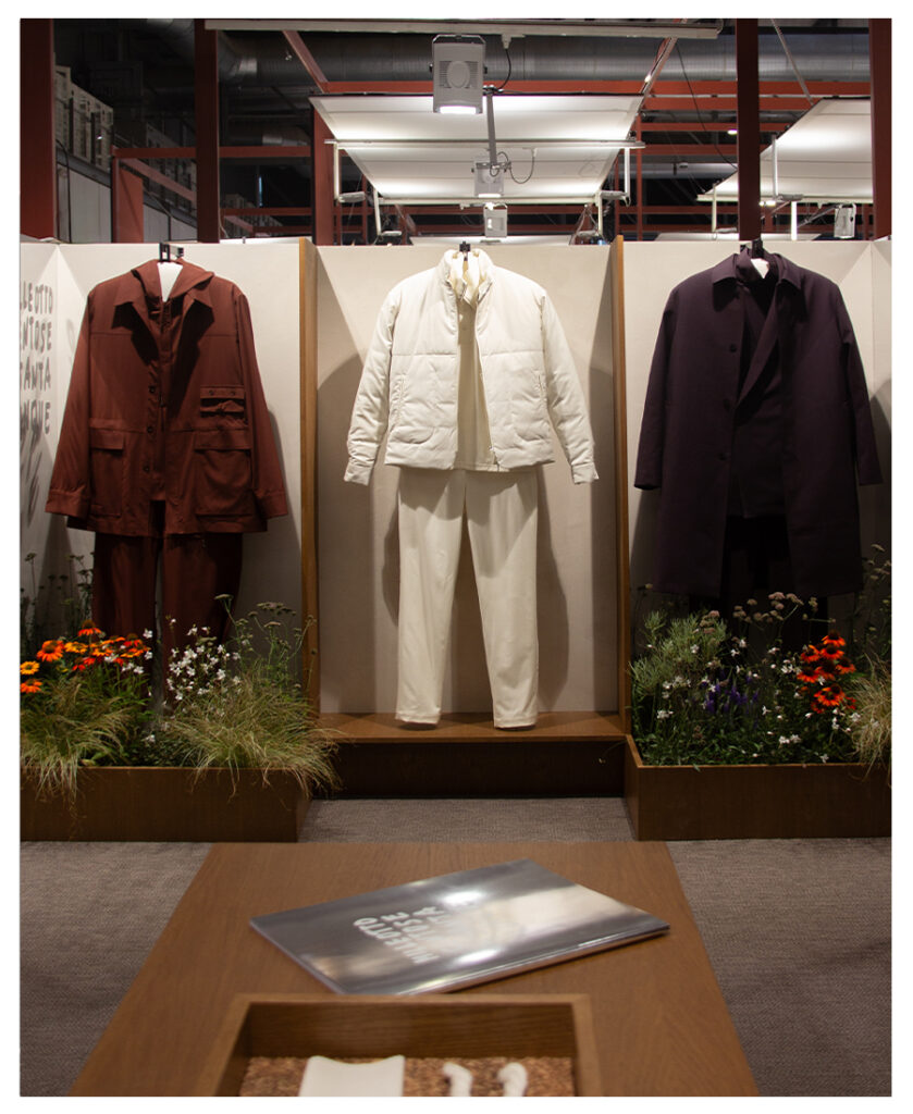 Three outfits on display: rust-brown jacket set, white padded jacket with trousers, and dark coat, among flowers