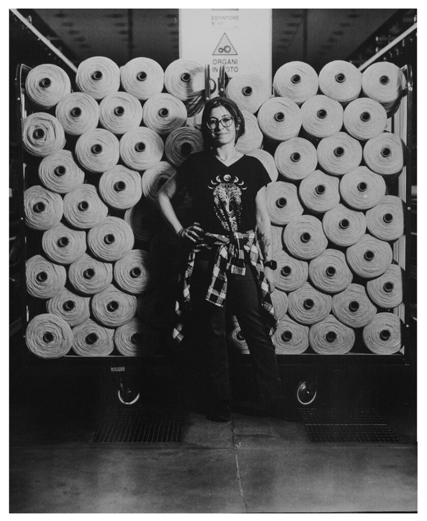 Woman with glasses stands smiling, hands on hips, in front of a large stack of wool yarn cones in a textile mill.