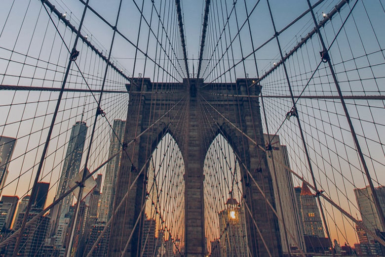 Stone tower of the Brooklyn Bridge seen through its web of steel cables at dusk, with skyscrapers behind