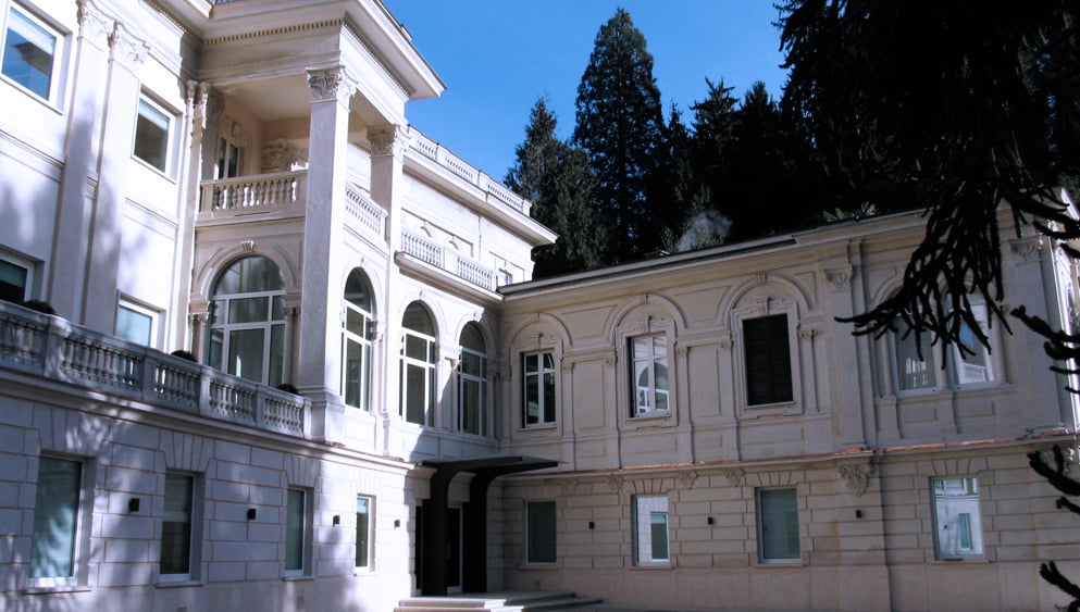 Cream-colored neoclassical villa with arched windows, balustrades and a columned balcony, beside tall evergreen trees