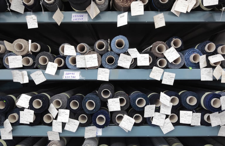 Rolls of blue, grey and black fabric with paper tags stacked on metal warehouse shelves