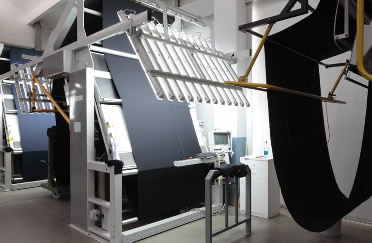 Dark fabric stretched over an inspection machine with lights, beside a control station, in a textile workshop
