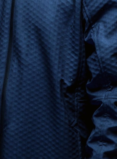 Close-up of a navy blue jacket with a quilted diamond texture, a vertical zip and a pocket opening.