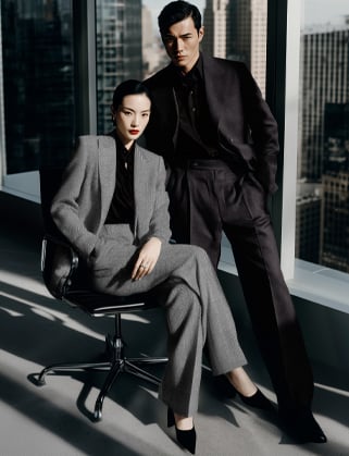 Woman in a grey suit seated on an office chair beside a standing man in dark attire, by a skyline window