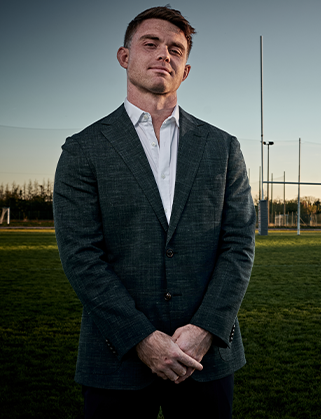 Man in a dark textured wool-look blazer and white open-collar shirt stands on a rugby pitch at dusk