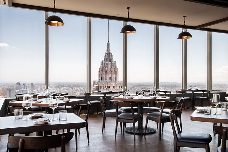 Restaurant dining room with wooden tables and leather chairs, with a skyscraper spire seen through tall windows